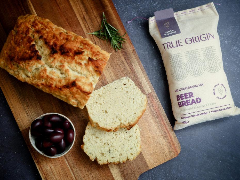 Loaf of Olive & Rosemary Beer Bread with slices on a wooden board next to a package of True Origin beer bread..