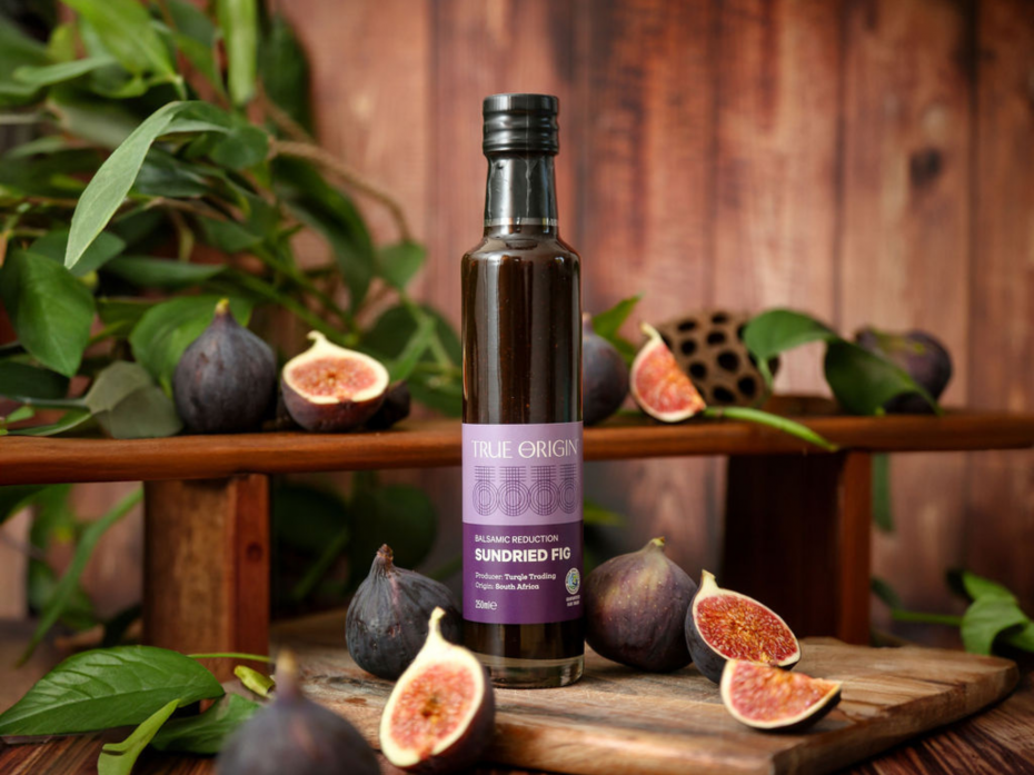 Purple bottle of True Origin Sundried Fig Balsamic Reduction on a wooden chopping board surrounded by figs and some plants in the background..
