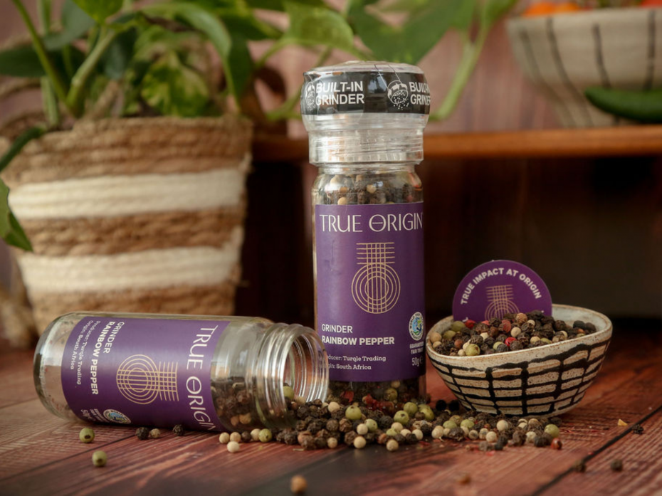 True Origin Rainbow Pepper grinder with open lid beside a bowl of rainbow peppercorns on a wooden surface